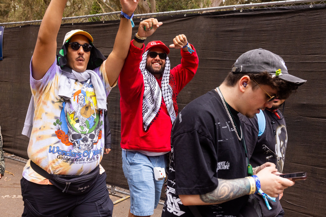 Three young men walk outdoors, one in a tie-dye shirt and sunglasses raising his arms, another in a red hoodie smiling, and the third focused on his phone.